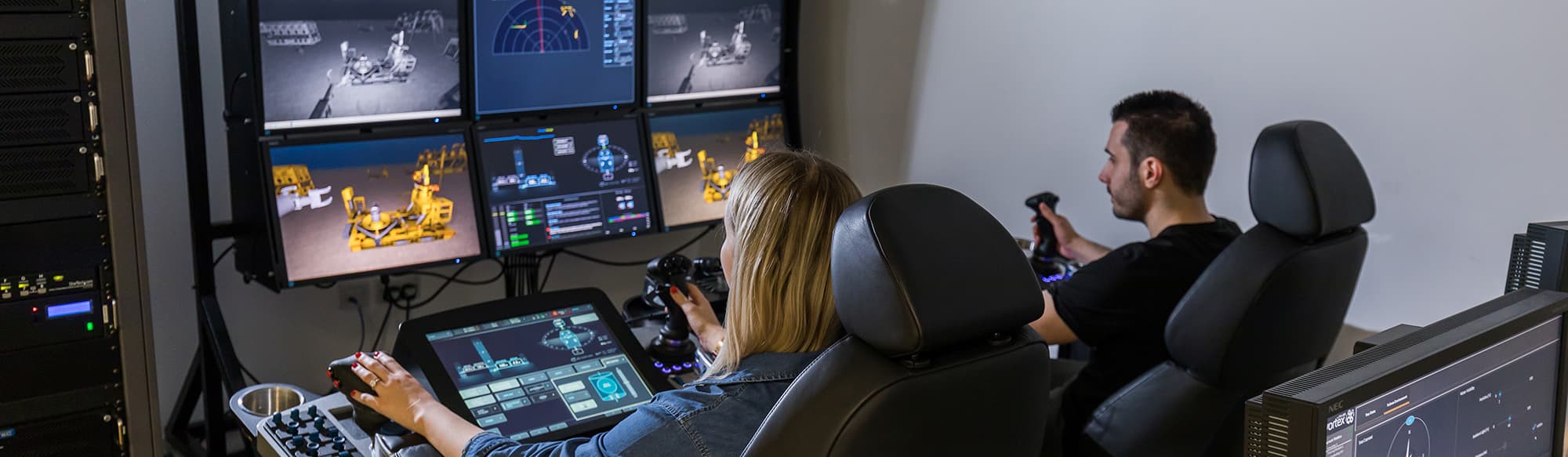 ROVOP staff using the ROVOP Academy training simulator. Two people using joysticks and touchscreens sit in front of a bank of monitors