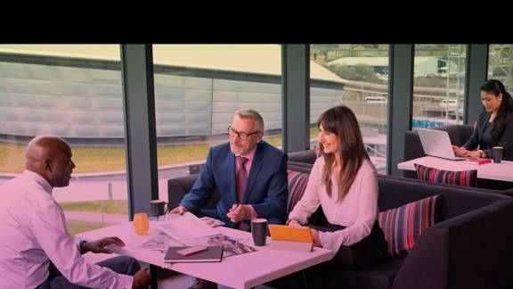 Three business people having a meeting round a table that is strewn with documents and notebooks. Through the window can be seen the Glasgow venue, the OVO Hydro