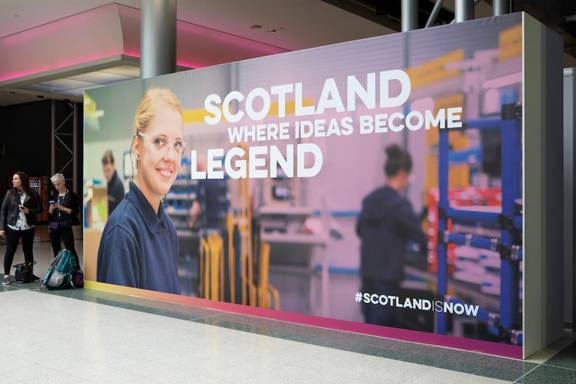 Two people at a conference stand beside a billboard-sized poster with the headline: Scotland - where ideas become legend