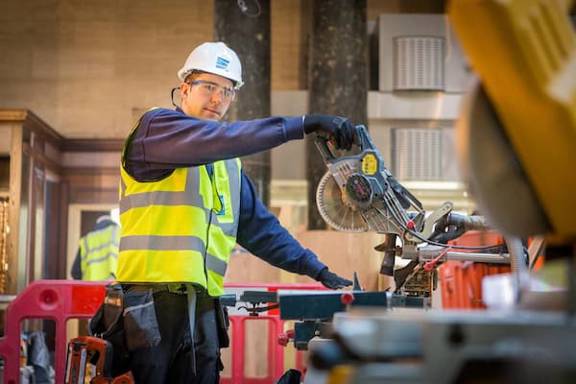 A worker wearing hi-viz and safety equipment uses a circular saw