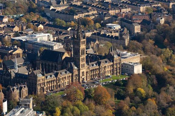 Anaerial view of Glasgow University. The main building is in the centre, its gothic facade glowing in Autumn sunlight. In the foreground, the trees of Kelvingrove Park are just beginning to turn.